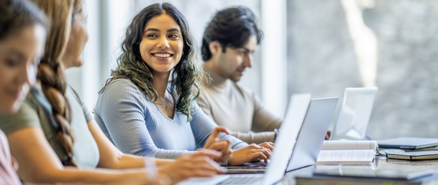 A small group of four mature adult students sit together at a long desk as they study with one another. They have text books and laptops out in front of them as they take notes and talk amongst themselves.