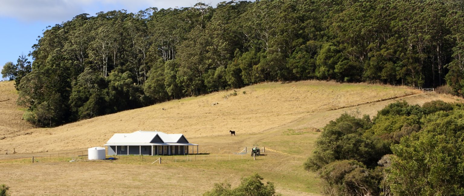 A modern farm house on a hill side with trees surrounding it, a water tank, horse and a tractor are in the paddock.